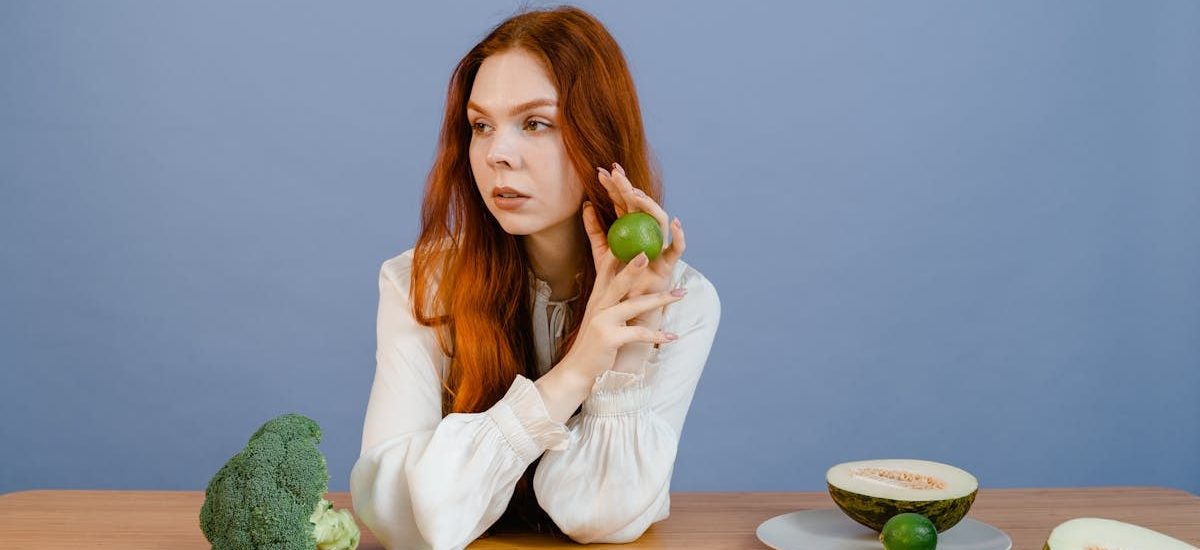 Caucasian woman sitting at table holding lime with broccoli and melon.