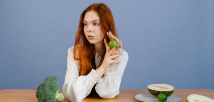Caucasian woman sitting at table holding lime with broccoli and melon.
